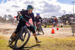 Mountain biker cornering hard on a dusty dirt track with cones and spectators behind