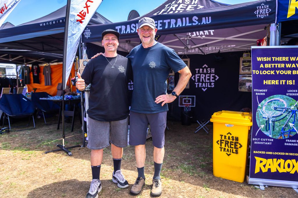 Dan and Tom from Trash Free Trails standing in front of their stand beside a yellow bin