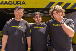 Three men in Magura shirts laughing together at the Magura stand